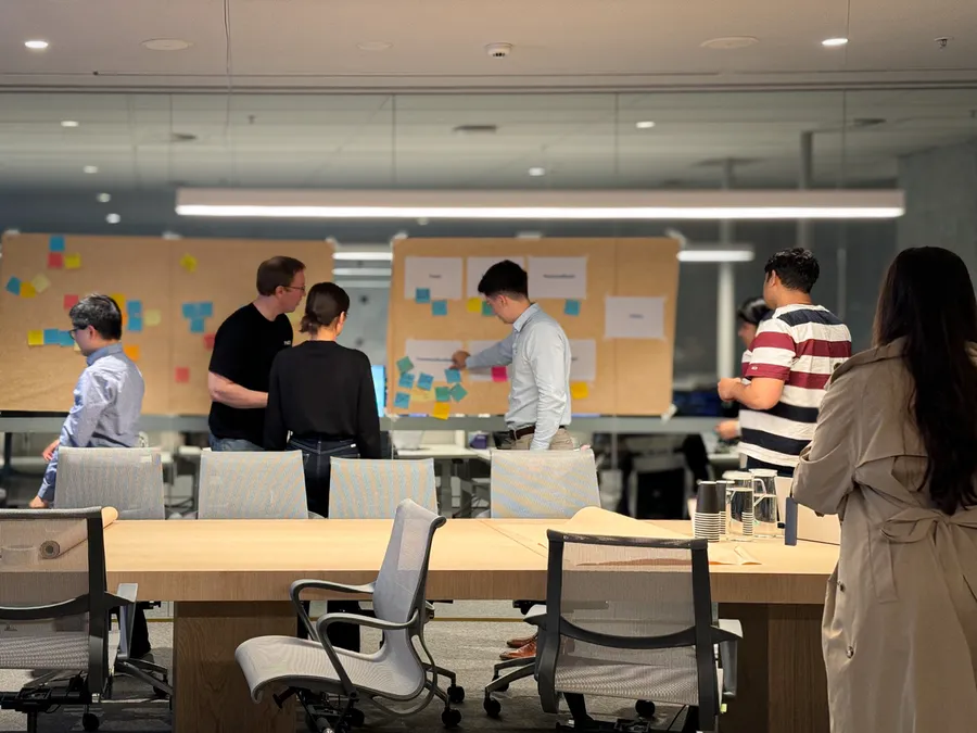 A team gathered at pinboards of sticky notes across an open office floor, seen from the far end of a meeting table.